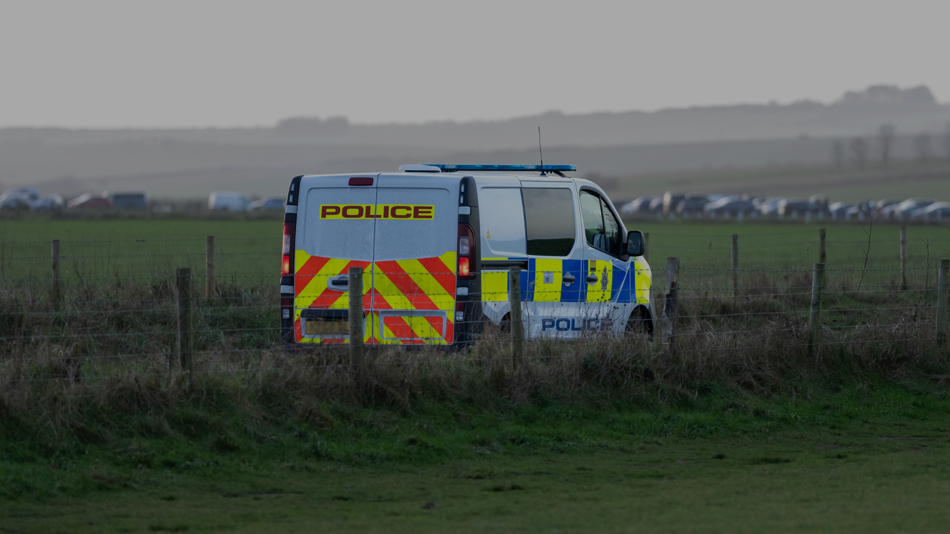 A police van parked in a field behind a wire fence, with a row of vehicles visible in the distance.