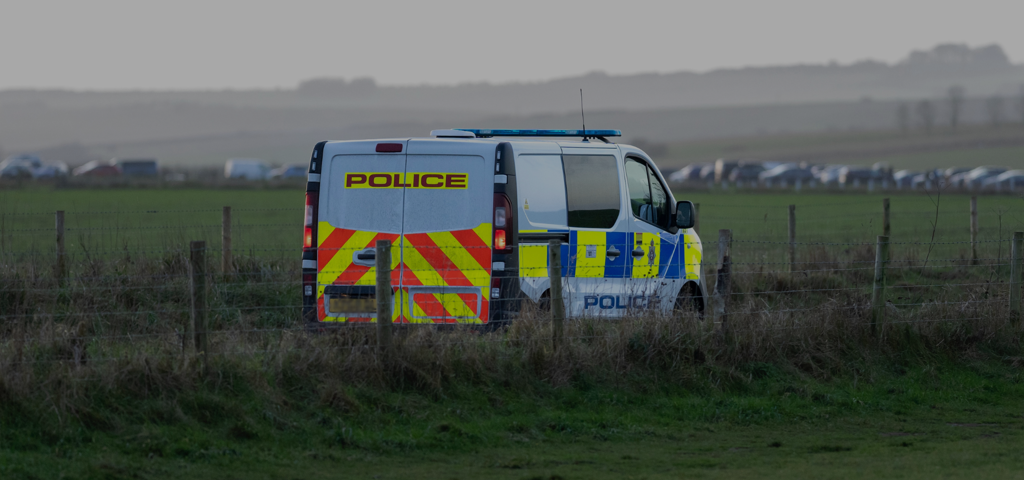 A police van parked in a field behind a wire fence, with a row of vehicles visible in the distance.