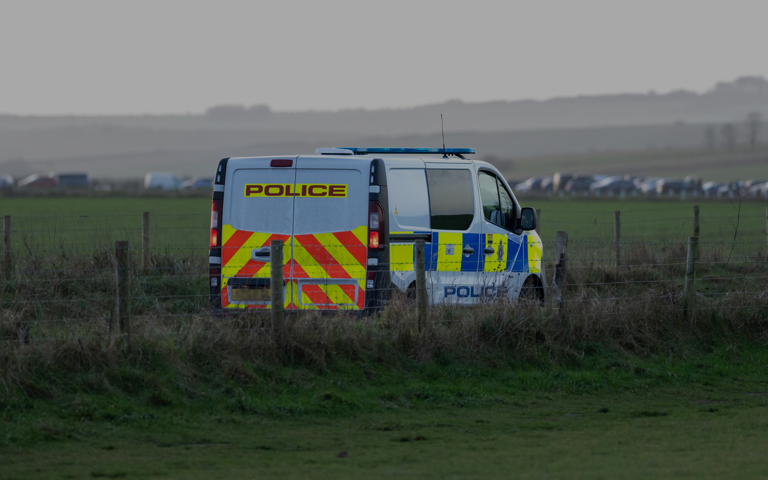 A police van parked in a field behind a wire fence, with a row of vehicles visible in the distance.