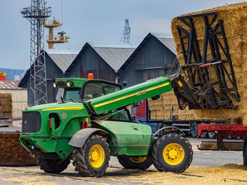 Green telehandler in action, unloading straw bales from a red lorry on a rural site.