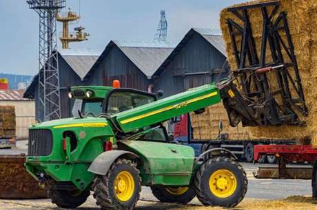 Green telehandler in action, unloading straw bales from a red lorry on a rural site.