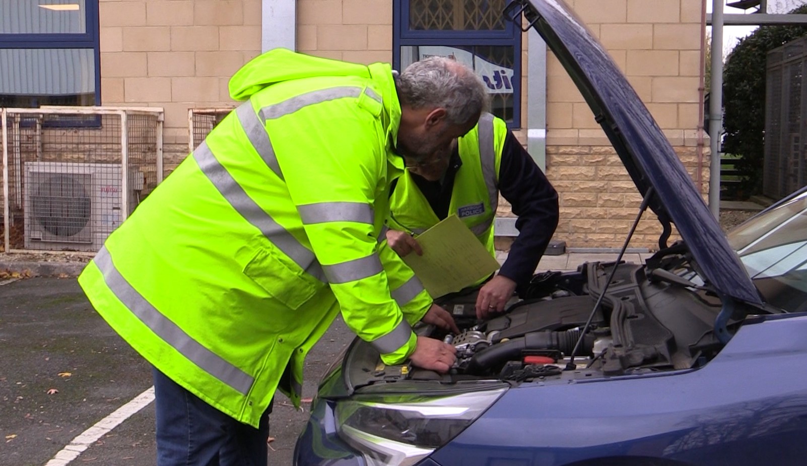 Two gentlemen examining the engine bay of a blue car during a motor vehicle inspection.