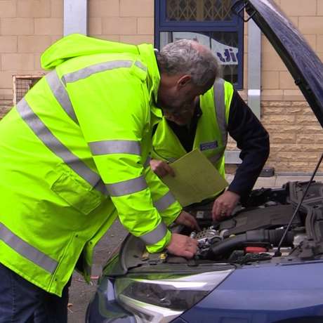 Two gentlemen examining the engine bay of a blue car during a motor vehicle inspection.