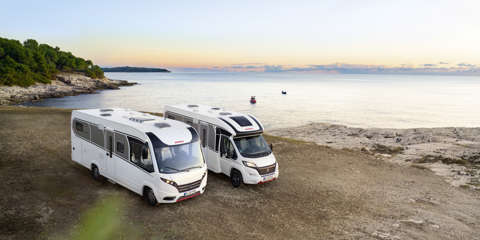 Two motorhomes parked near a coastal view as the sun sets over the horizon.