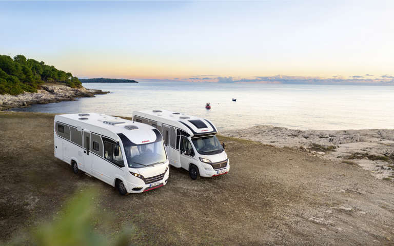 Two motorhomes parked near a coastal view as the sun sets over the horizon.