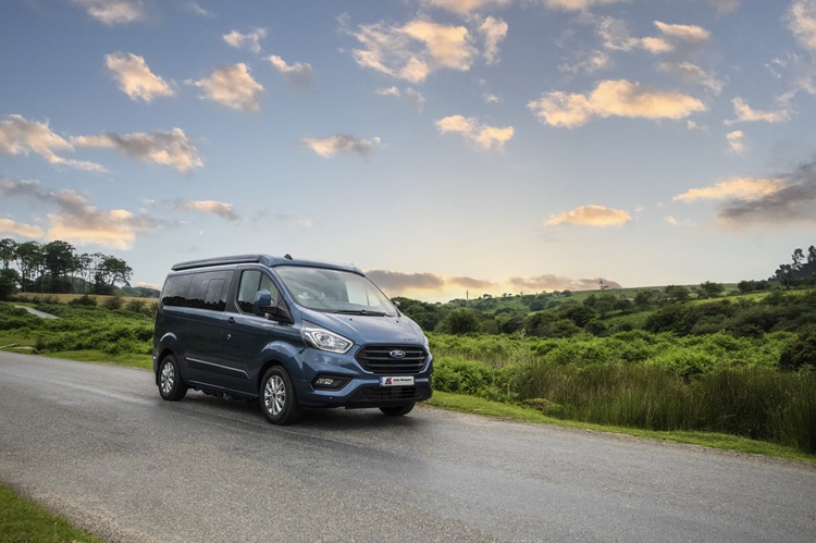 A blue campervan traveling along a scenic road in vibrant, green countryside.