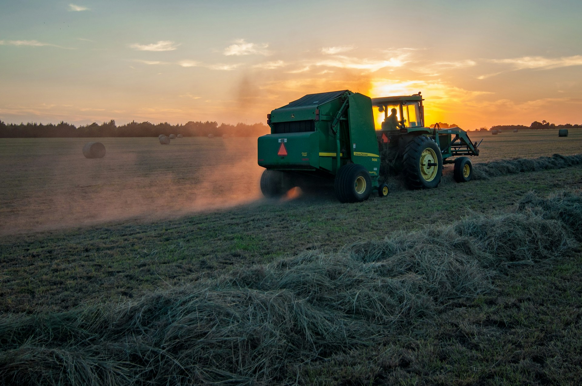 Evening harvest with green tractor and round baler silhouetted against the sunset.