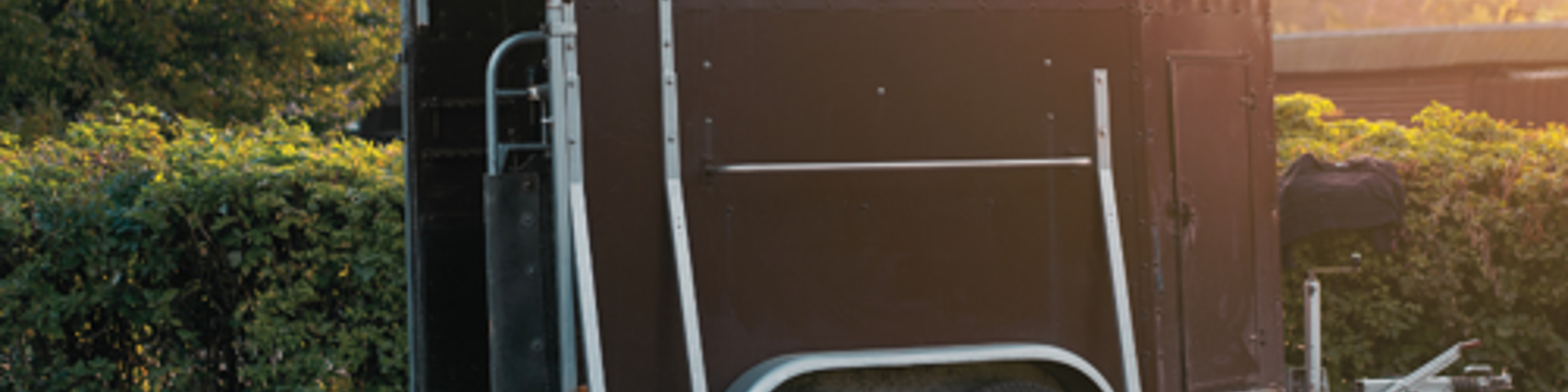 A brown horse trailer featuring a white roof and lowered ramp, illuminated by bright sunlight.