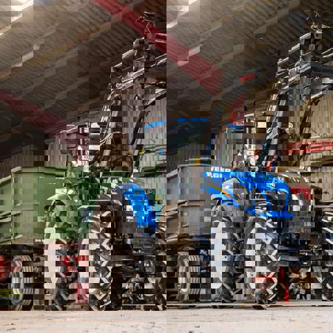 A blue utility loader tractor attached to a green trailer parked in a farmyard setting.