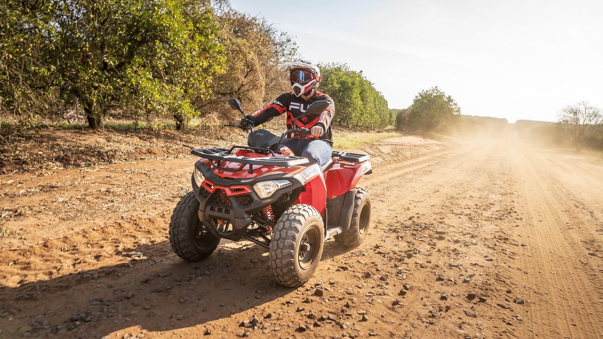 Stationary red ATV with rider on a dirt track, framed by a bright sun in the background.