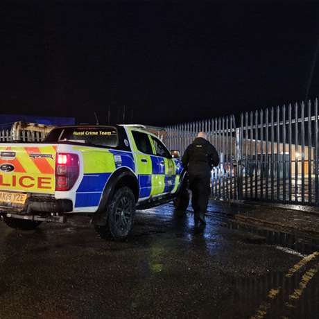 Police vehicle parked with an officer approaching locked gates at night.