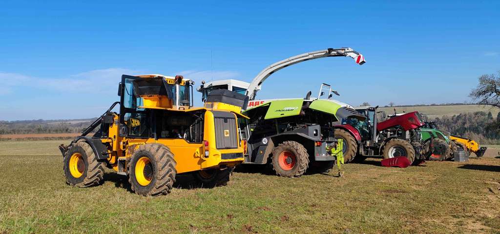 Line-up of farm machinery including JCB telehandler, CLAAS Jaguar forage harvester, tractors and vans on a farm field under a clear blue sky.