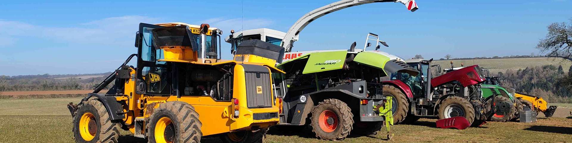 Line-up of farm machinery including JCB telehandler, CLAAS Jaguar forage harvester, tractors and vans on a farm field under a clear blue sky.