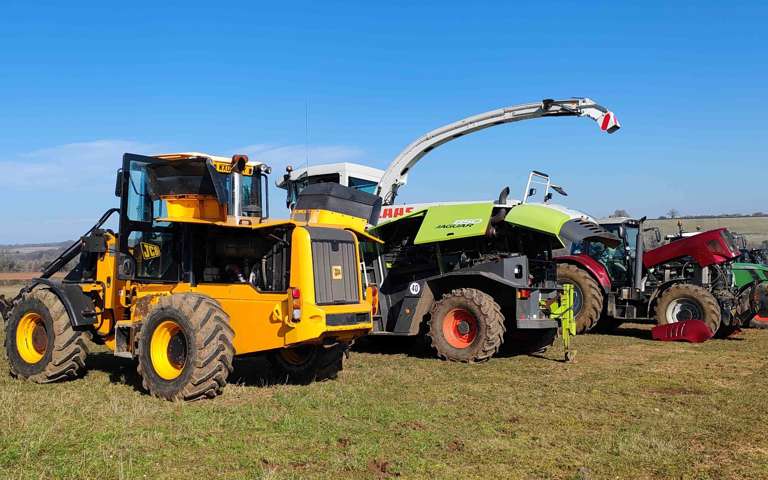 Line-up of farm machinery including JCB telehandler, CLAAS Jaguar forage harvester, tractors and vans on a farm field under a clear blue sky.