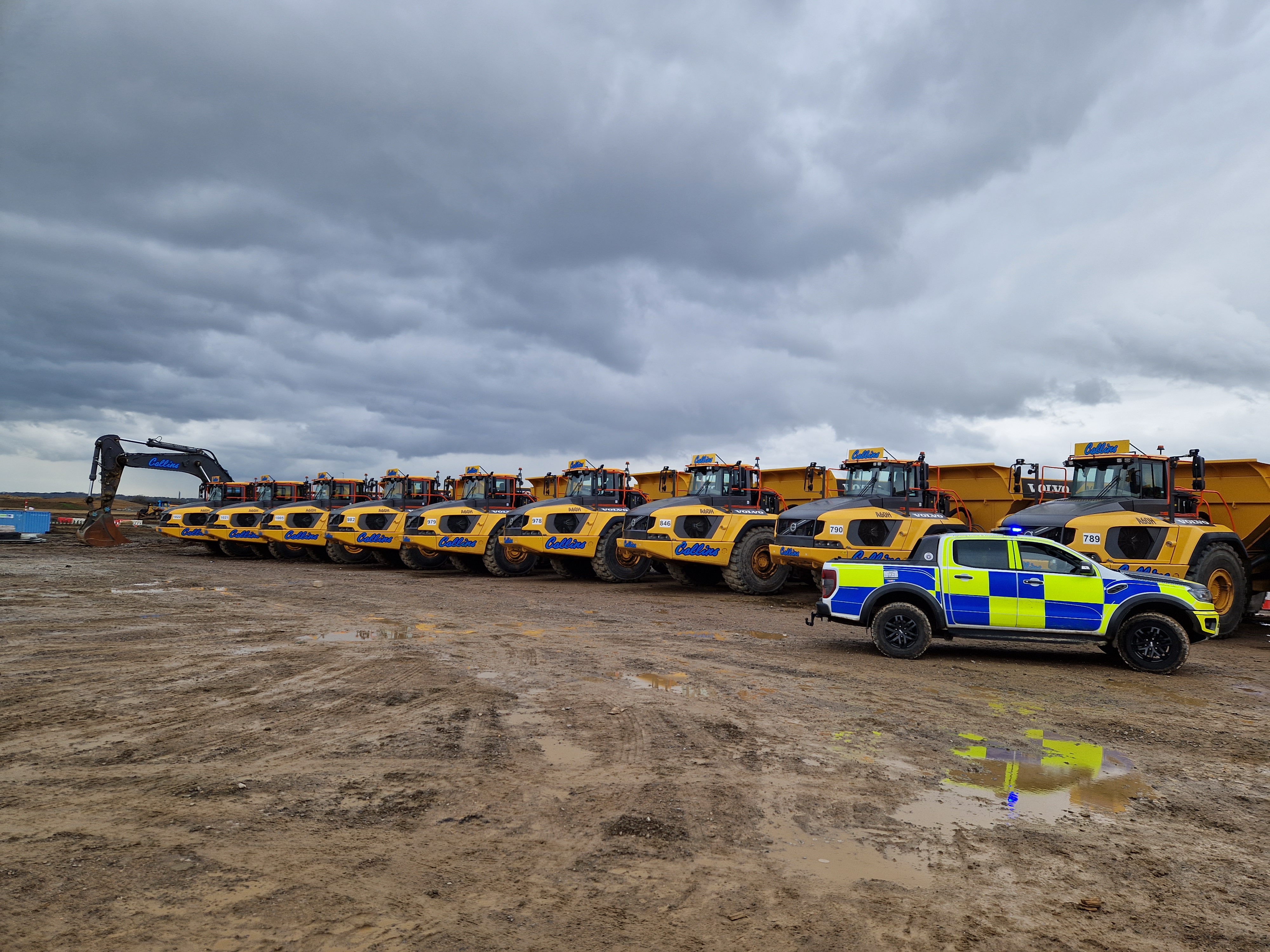 A police car parked to the right of a line of yellow off-road dumpers on a construction site.