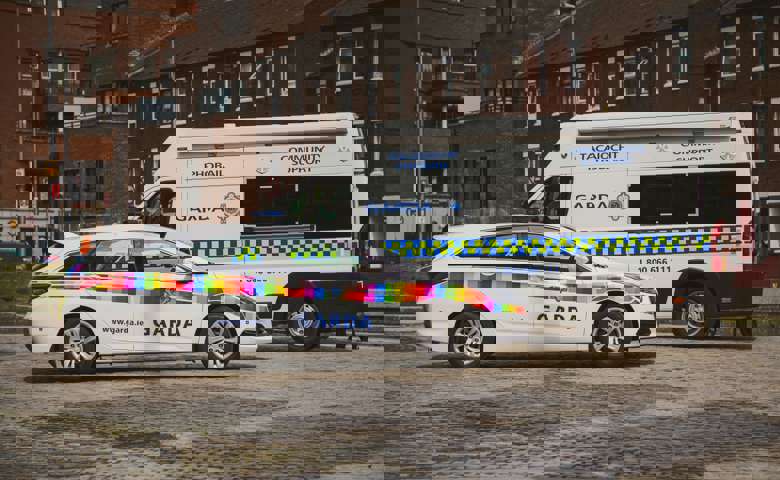 Rainbow flag-branded Garda car and a Garda community support van parked on a cobblestone street.