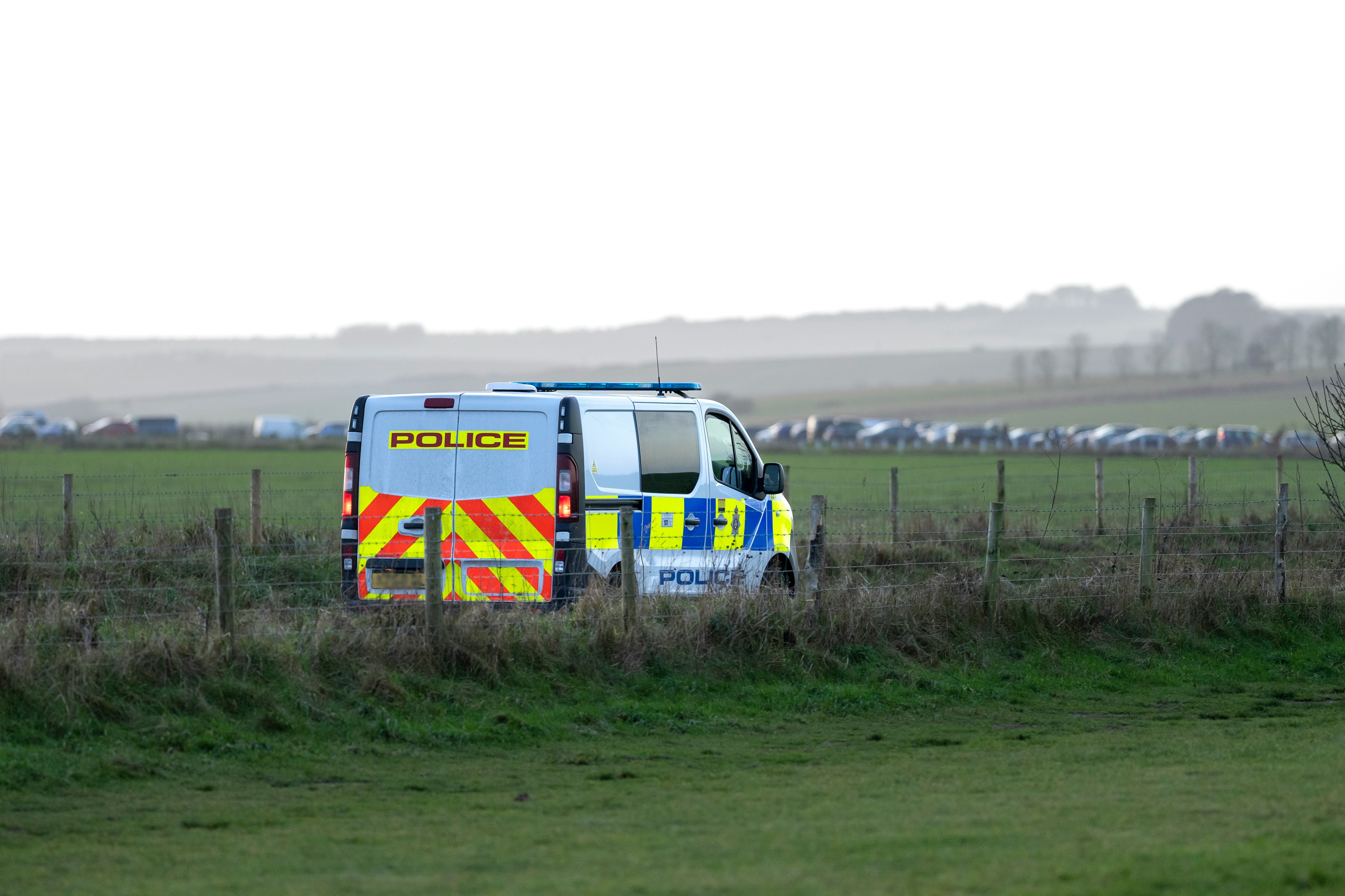 A police van parked in a field behind a wire fence, with a row of vehicles visible in the distance.