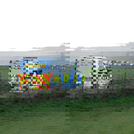 A police van parked in a field behind a wire fence, with a row of vehicles visible in the distance.