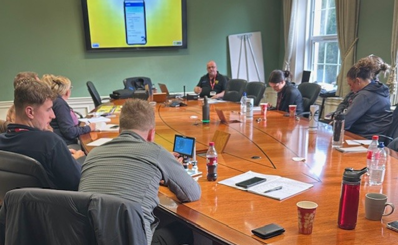Police officers gathered around a large wooden table, watching a PowerPoint presentation on a large screen during a VIN CHIP training session.