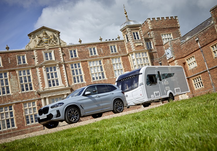 Silver car with a white caravan, stationary in front of an elegant stately home.