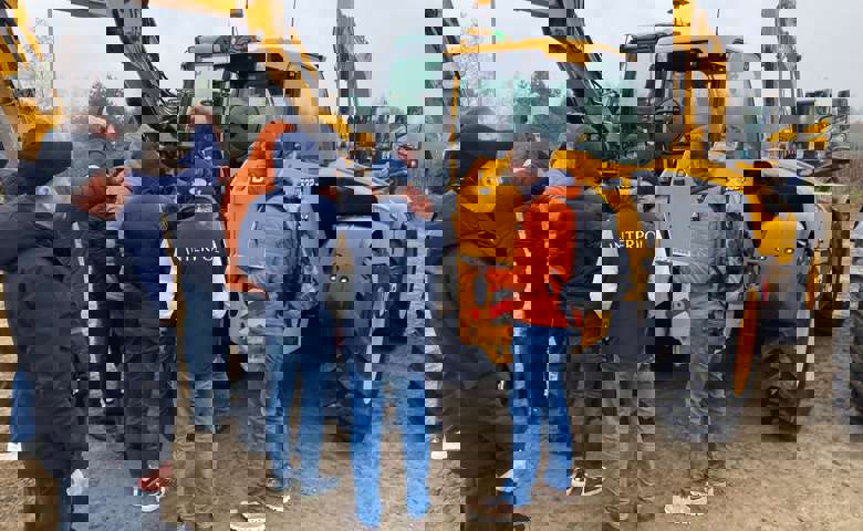 A group of students engaged in training, with JCBs and a rural landscape in the background.