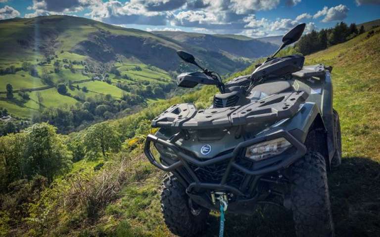 Green CFMOTO ATV parked on rolling hills under dramatic cloudy sky with sunbeams.