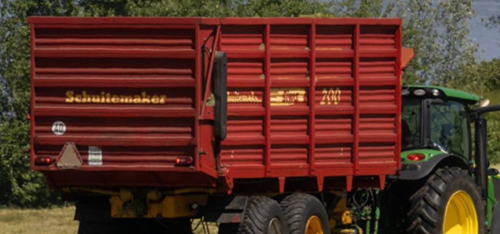 A green tractor pulls a bright red-orange agricultural trailer in a field by a lake, with a sailboat visible in the distance.