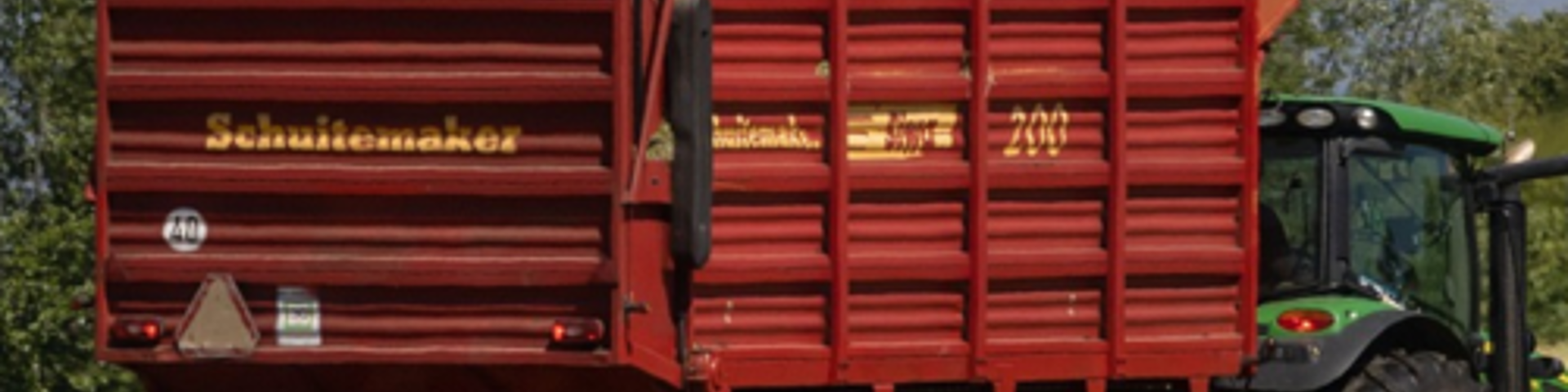 A green tractor pulls a bright red-orange agricultural trailer in a field by a lake, with a sailboat visible in the distance.