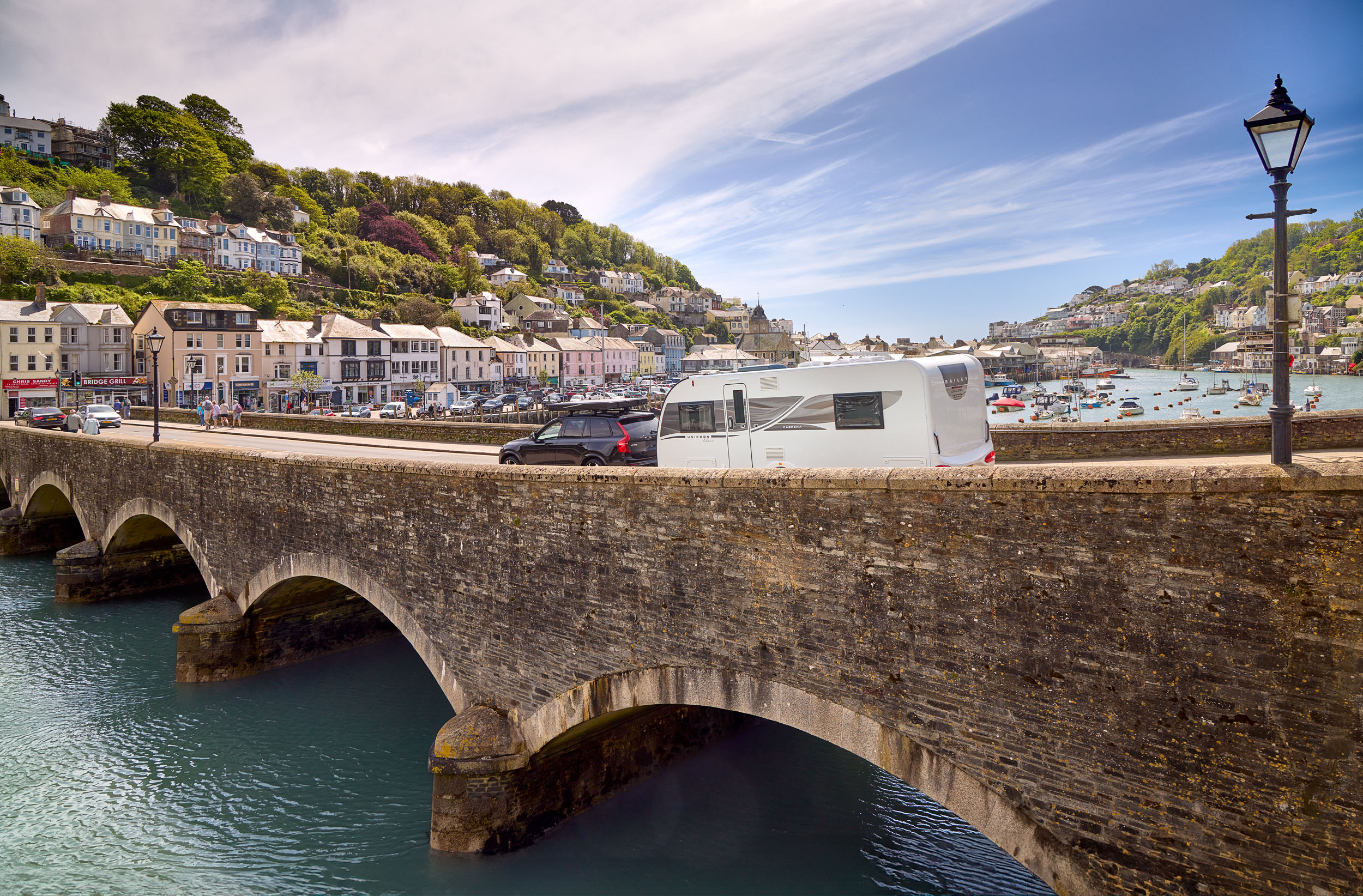 A family caravan is towed by a car over a scenic arched bridge, with the ocean in the background.