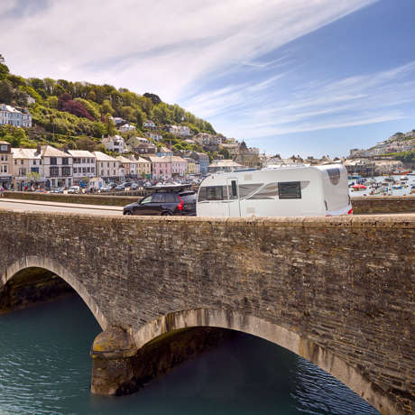 A family caravan is towed by a car over a scenic arched bridge, with the ocean in the background.
