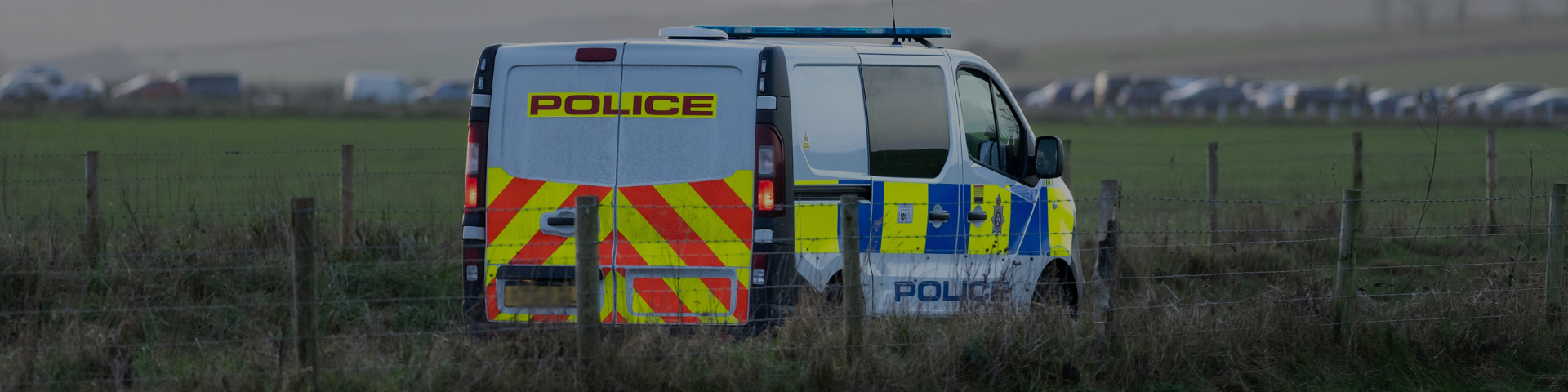 A police van parked in a field behind a wire fence, with a row of vehicles visible in the distance.