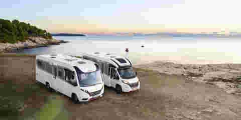 Two motorhomes parked near a coastal view as the sun sets over the horizon.