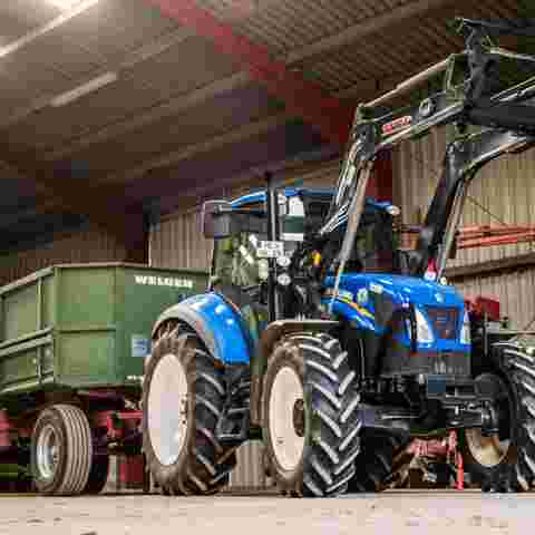 A blue utility loader tractor attached to a green trailer parked in a farmyard setting.