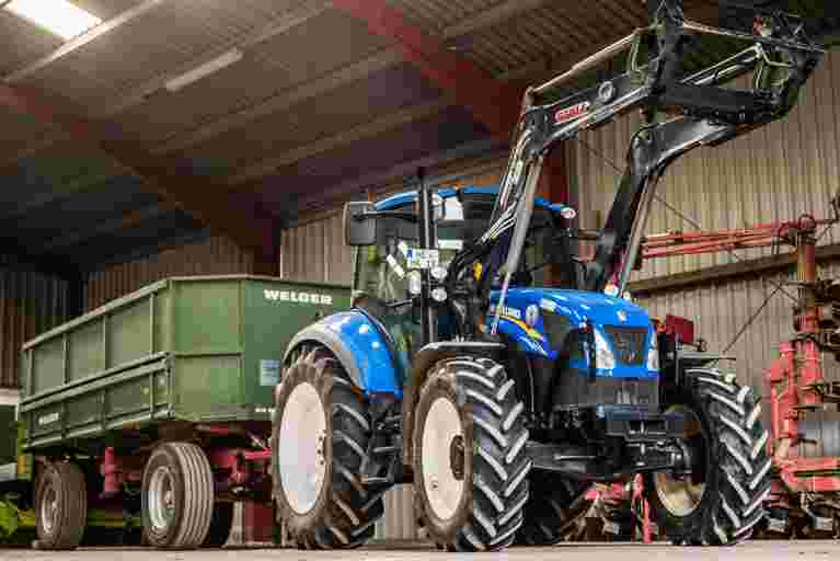 A blue utility loader tractor attached to a green trailer parked in a farmyard setting.