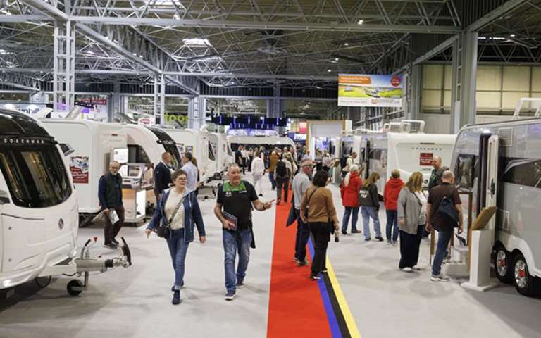 Visitors walking along a red-carpeted aisle at the Motorhome & Caravan Show, with caravans and motorhomes on either side
