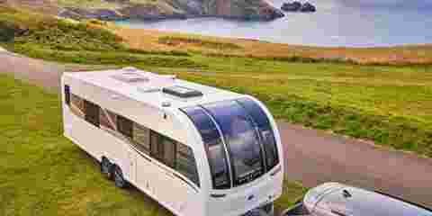 A silver car is pulling a twin axle caravan along a cliff road, overlooking a beautiful ocean landscape