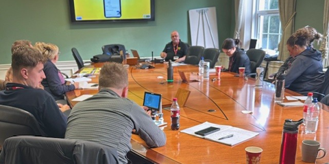 Police officers gathered around a large wooden table, watching a PowerPoint presentation on a large screen during a VIN CHIP training session.