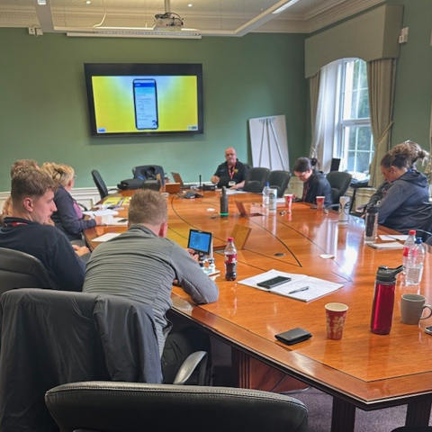 Police officers gathered around a large wooden table, watching a PowerPoint presentation on a large screen during a VIN CHIP training session.