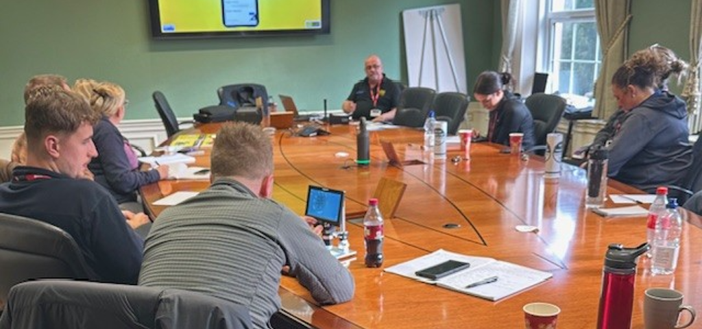Police officers gathered around a large wooden table, watching a PowerPoint presentation on a large screen during a VIN CHIP training session.