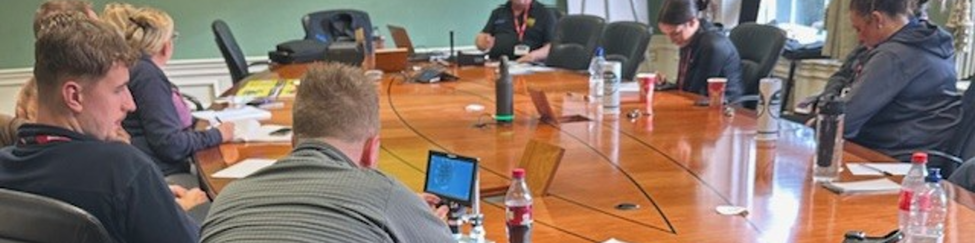 Police officers gathered around a large wooden table, watching a PowerPoint presentation on a large screen during a VIN CHIP training session.
