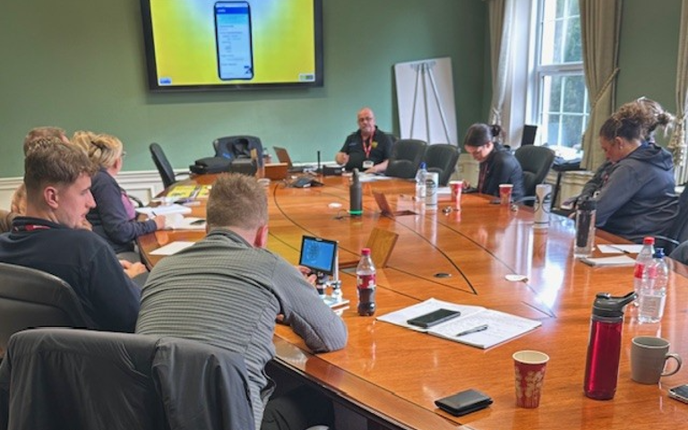 Police officers gathered around a large wooden table, watching a PowerPoint presentation on a large screen during a VIN CHIP training session.