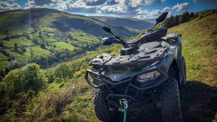 Green CFMOTO ATV parked on rolling hills under dramatic cloudy sky with sunbeams.