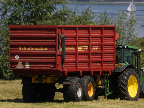 A green tractor pulls a bright red-orange agricultural trailer in a field by a lake, with a sailboat visible in the distance.
