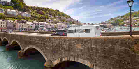  A family caravan is towed by a car over a scenic arched bridge, with the ocean in the background.