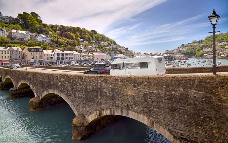  A family caravan is towed by a car over a scenic arched bridge, with the ocean in the background.